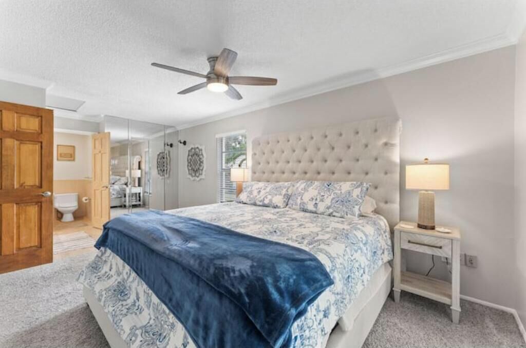 A bedroom with a bed, a ceiling fan, a bedside table with a lamp, and a door leading to the bathroom.