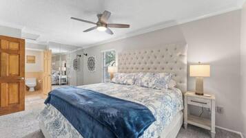 A bedroom with a bed, a ceiling fan, a bedside table with a lamp, and a door leading to the bathroom.