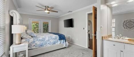 Un dormitorio con cama, mesita de noche, lámpara, ventilador de techo y televisor en la pared.