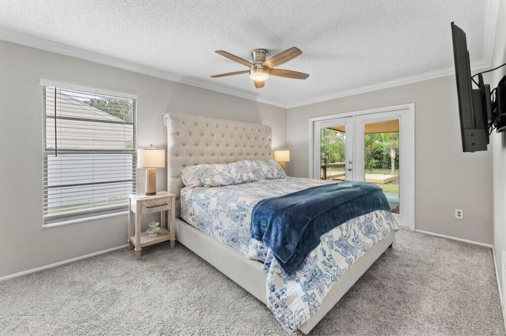 A bedroom with a bed, a ceiling fan, a nightstand, a lamp, and a view through a door to an outdoor area.