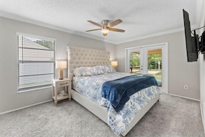 A bedroom with a bed, a ceiling fan, a nightstand, a lamp, and a view through a door to an outdoor area.