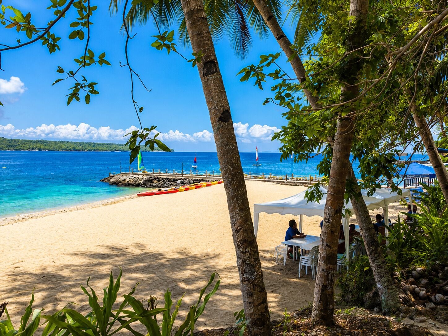 A beach with a clear view of the ocean, a pier with a sailboat, and a white tent with people seated.