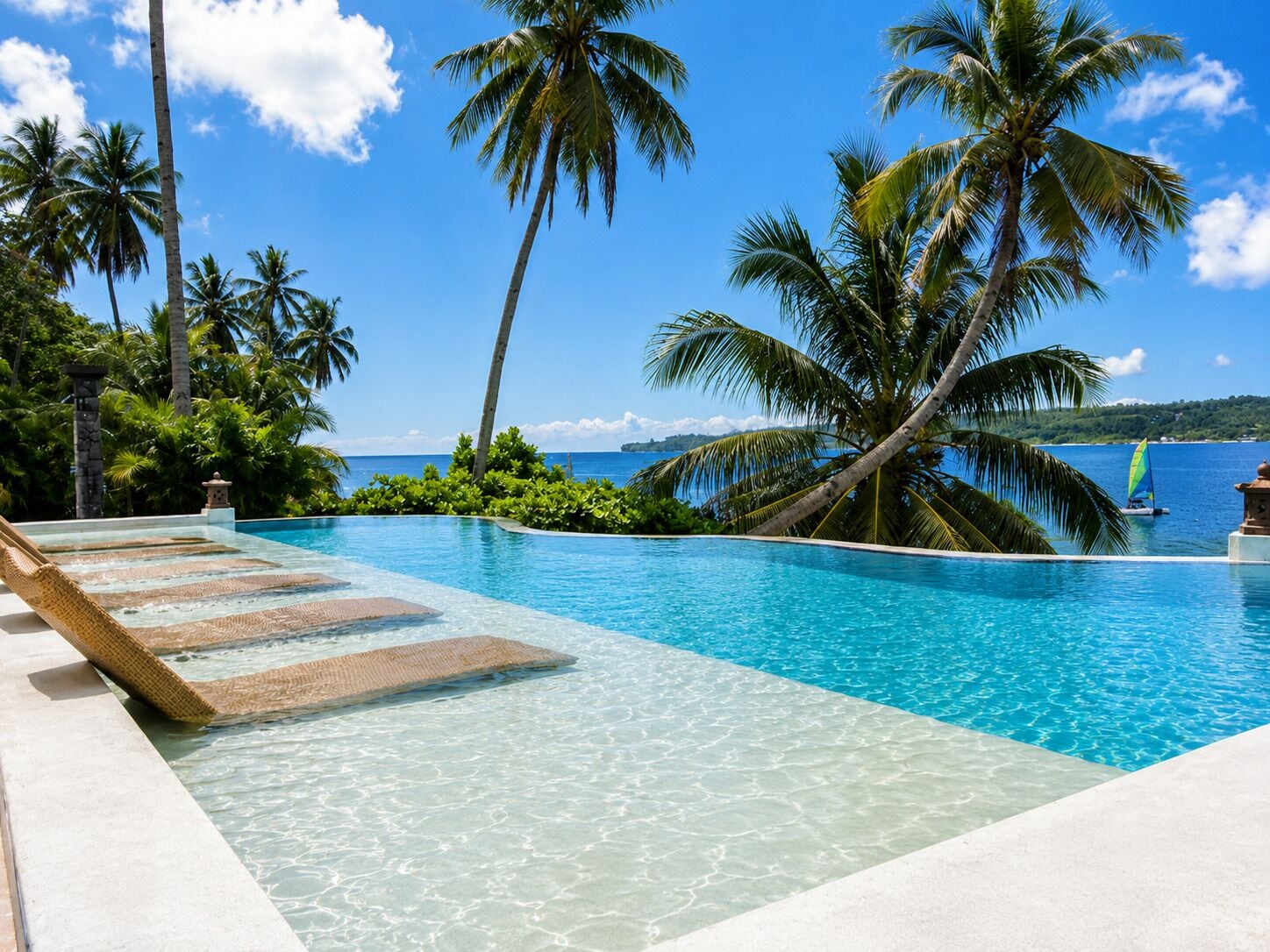 A clear blue pool with a wooden deck, surrounded by palm trees and a view of the ocean.