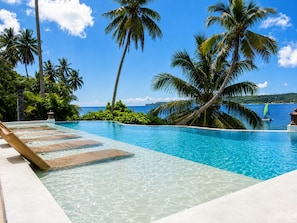 A clear blue pool with a wooden deck, surrounded by palm trees and a view of the ocean.