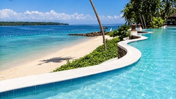A pool with a curved edge, a sandy beach, and a clear blue ocean.