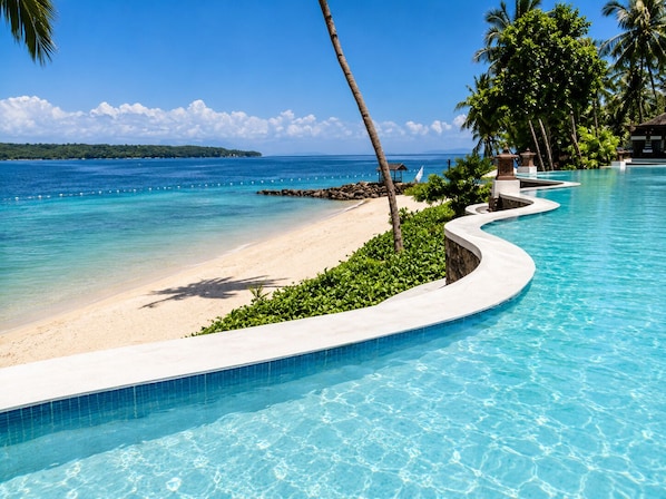 A pool with a curved edge, a sandy beach, and a clear blue ocean.