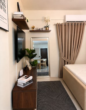 A bedroom with a bed, a desk with books, a mirror, a plant, and a window with curtains.
