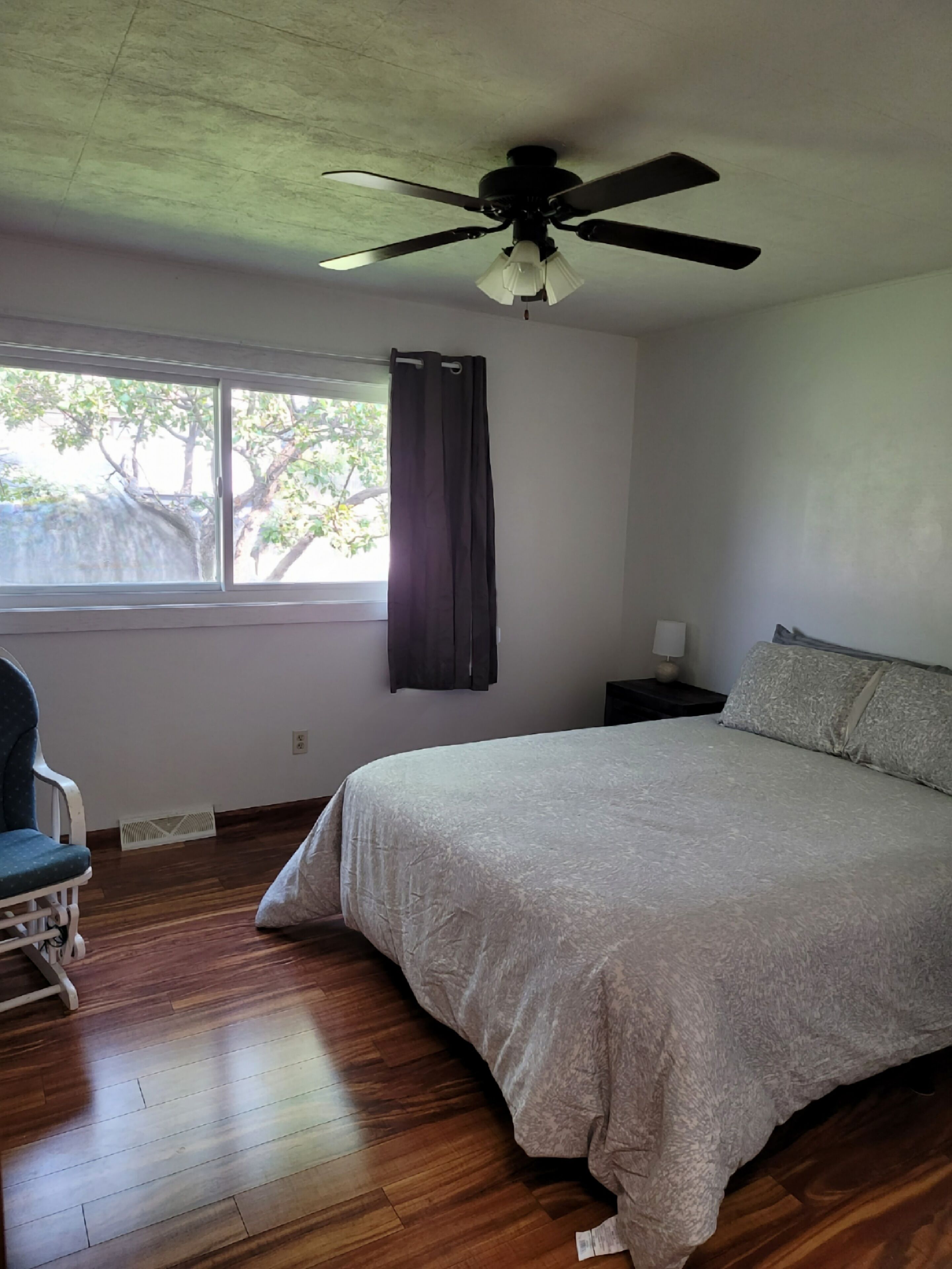 Un dormitorio con cama, ventilador de techo, ventana con cortinas, una silla y una mesita de noche con lámpara.