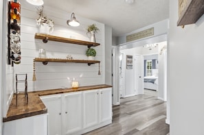 Cocina con armarios blancos, estantes de madera y una encimera. Se observan elementos decorativos y un colgante de techo.