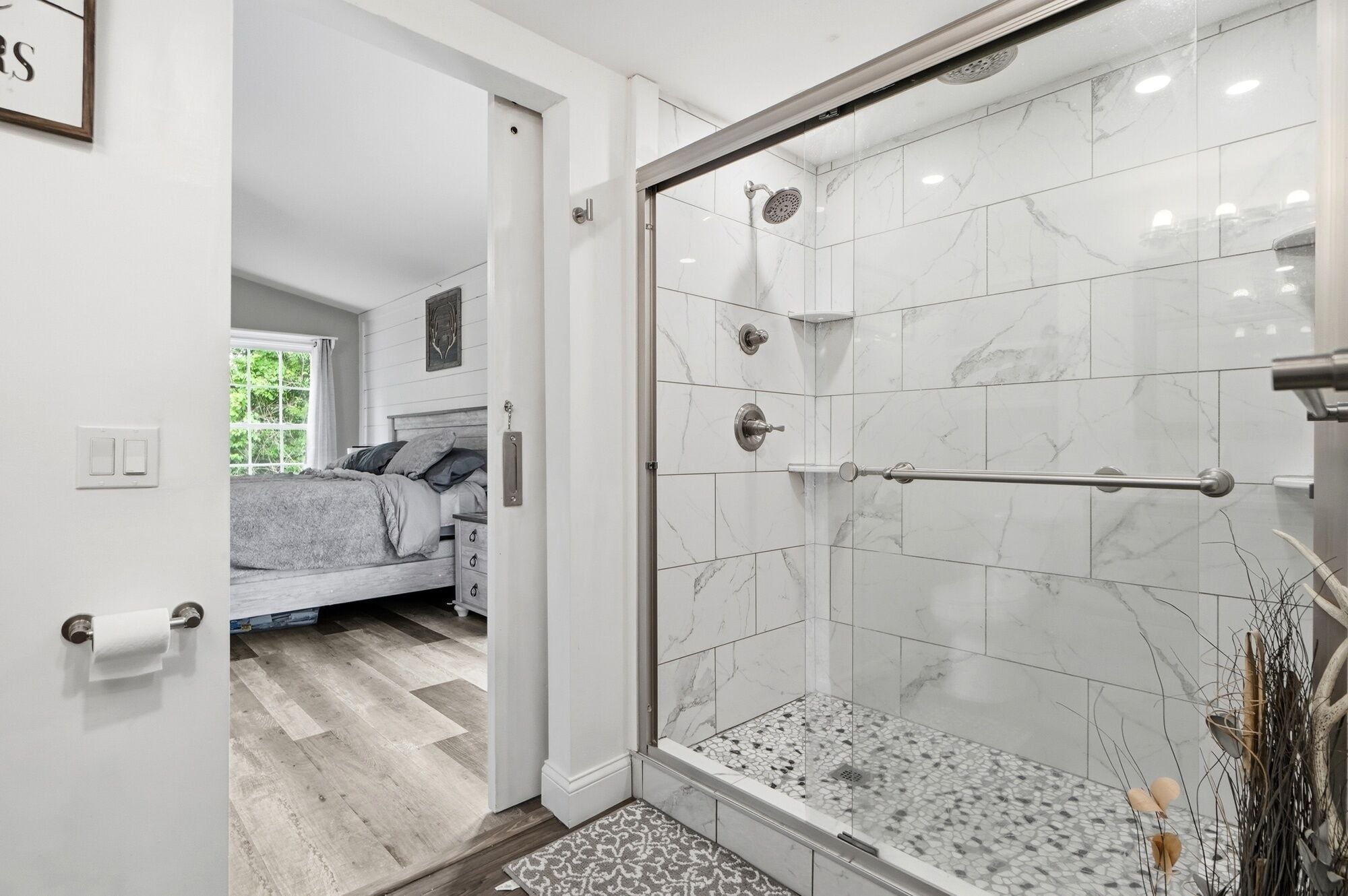 A modern bathroom with a glass-enclosed shower, marble tiles, and a visible bed in the adjacent room.