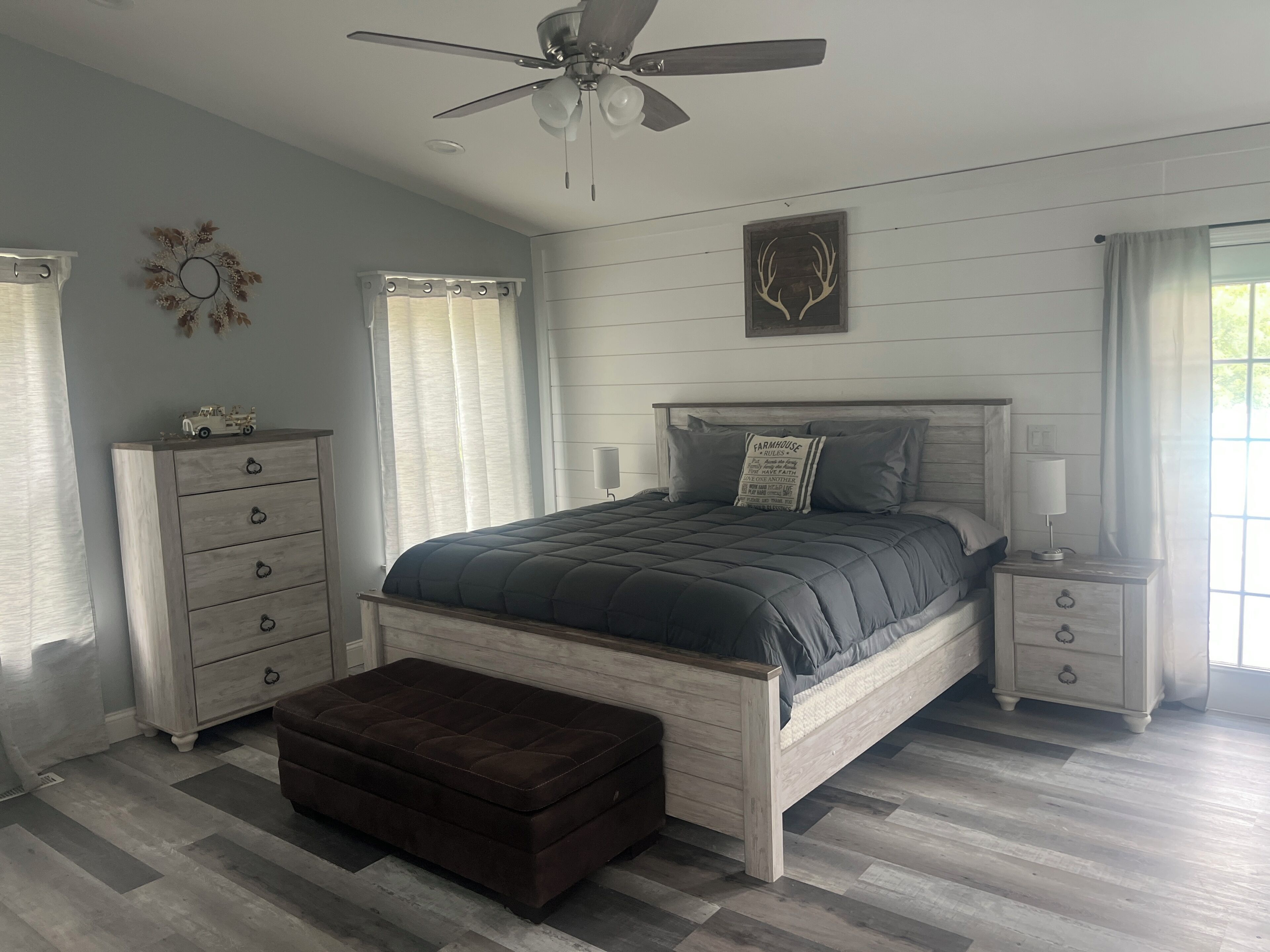 A bedroom with a bed, a chest of drawers, a nightstand, a ceiling fan, and a window with curtains.