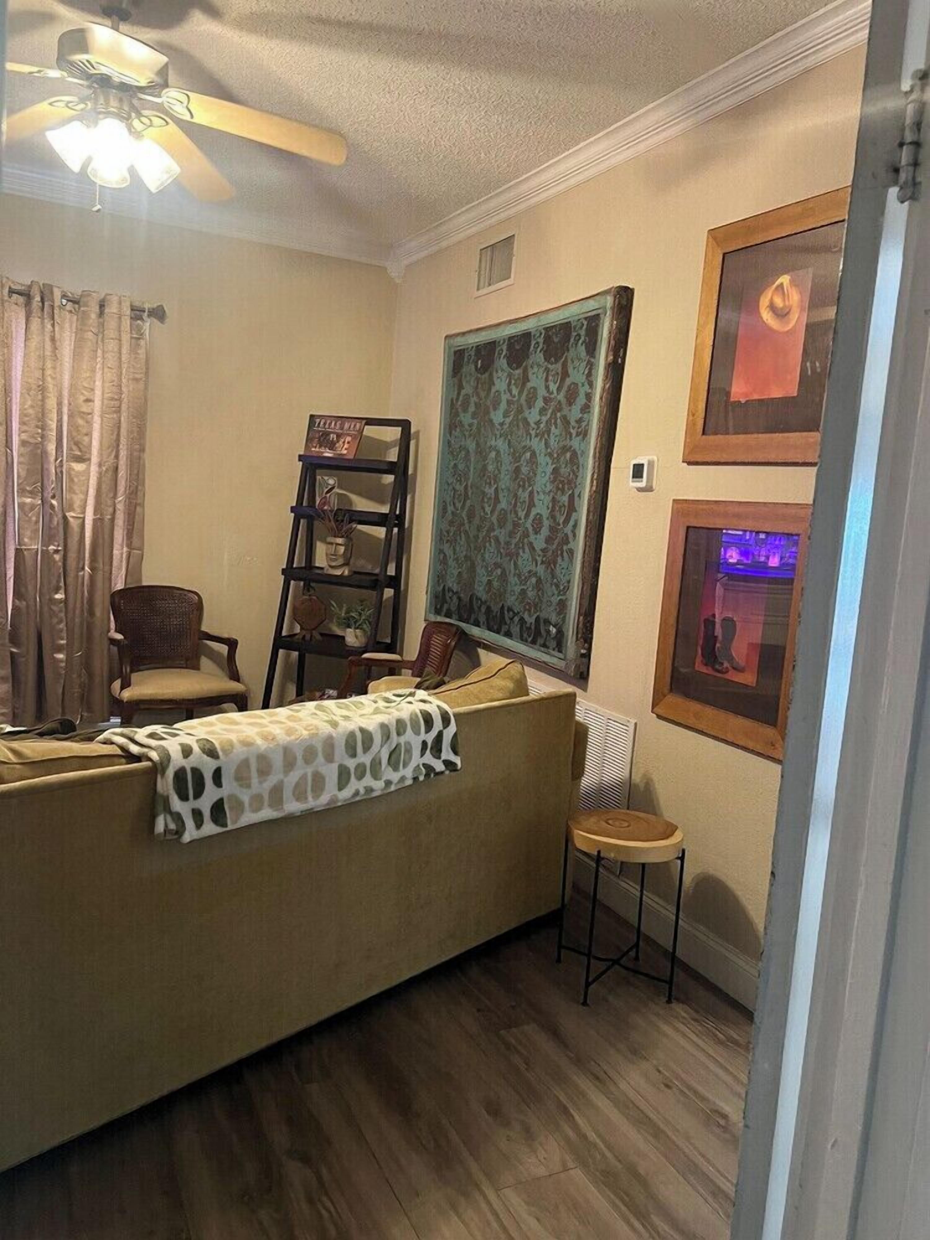 A room with a ceiling fan, a sofa, a chair, a small table, a bookshelf, and framed pictures on the wall.