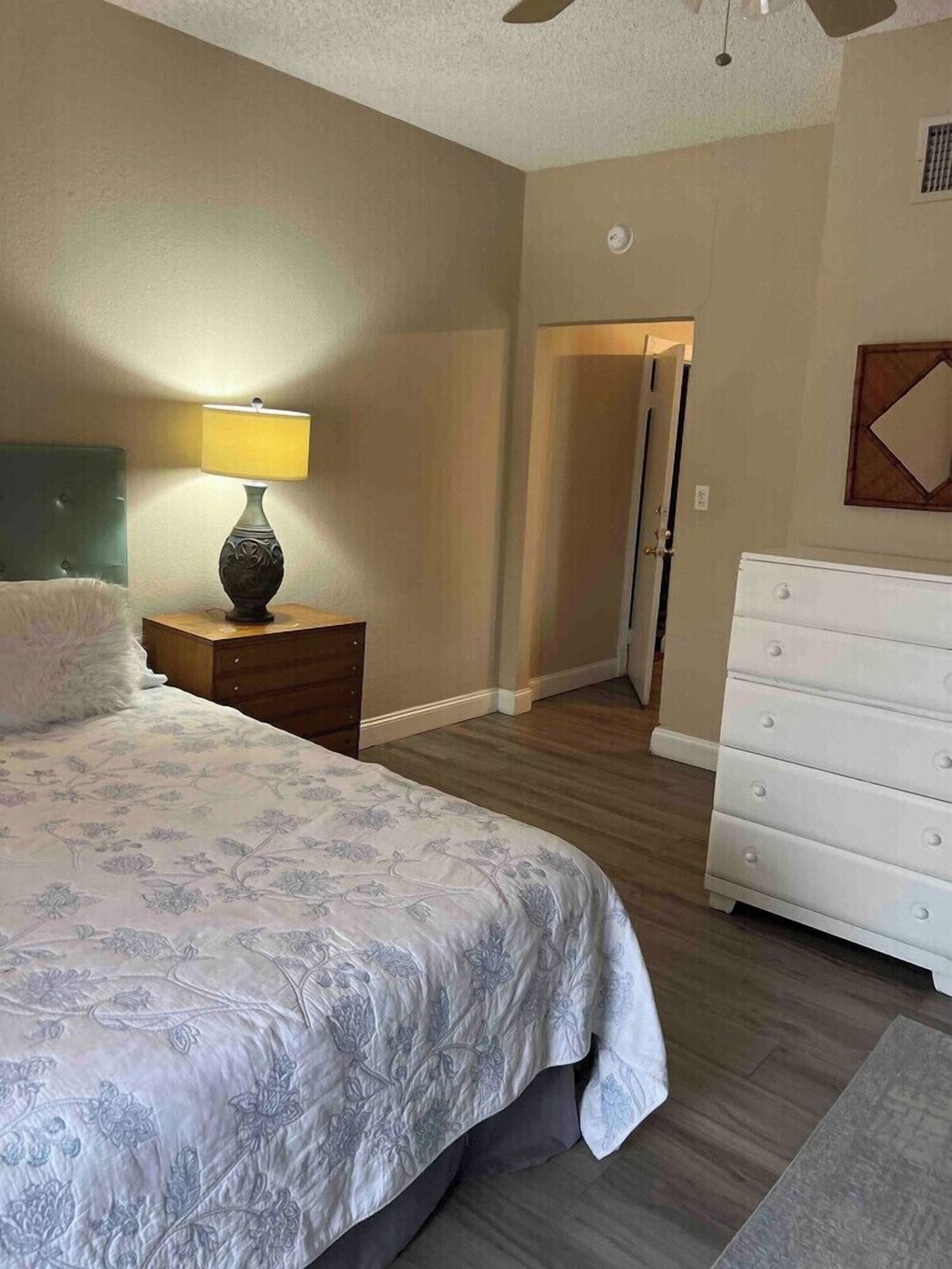 Un dormitorio con cama, mesita de noche con lámpara, cómoda y puerta que da a otra habitación.