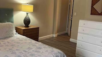 A bedroom with a bed, a nightstand with a lamp, a dresser, and a door leading to another room.