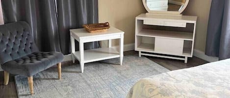 A bedroom with a bed, a chair, a small table, a mirror, and a dresser.