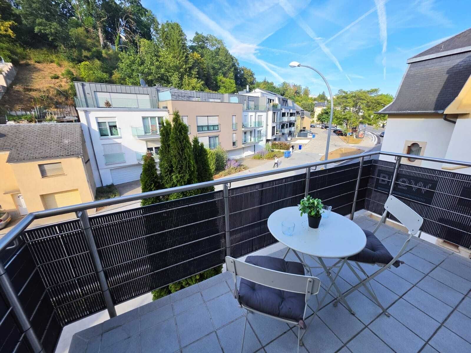 Een balkon met een tafel en stoelen, met uitzicht op een straat met moderne gebouwen en bomen.