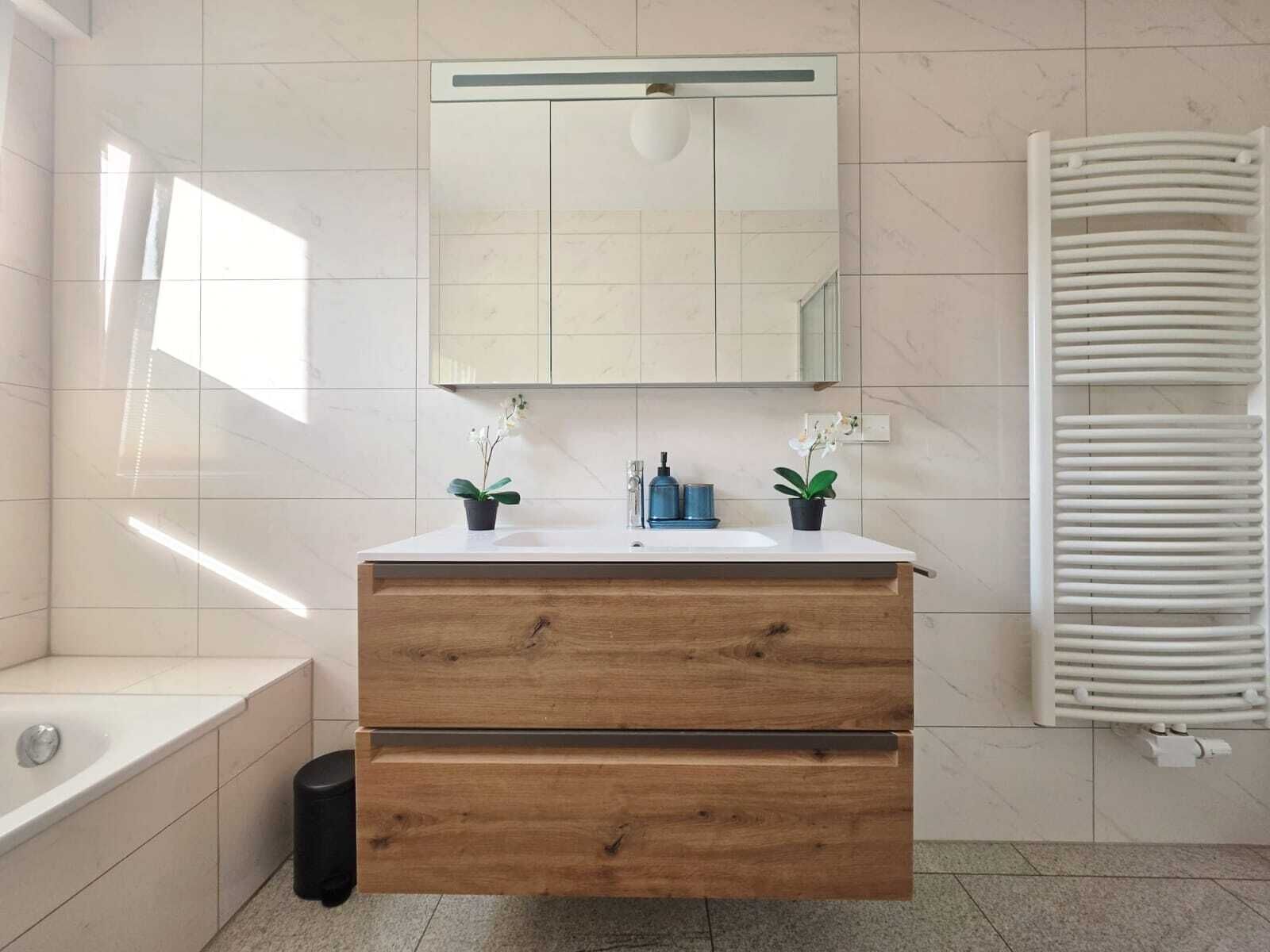 A bathroom with a wooden vanity, a mirror, two potted plants, a bathtub, and a radiator.
