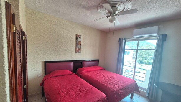 Una habitación con dos camas, un ventilador de techo, una ventana con cortinas y una puerta.