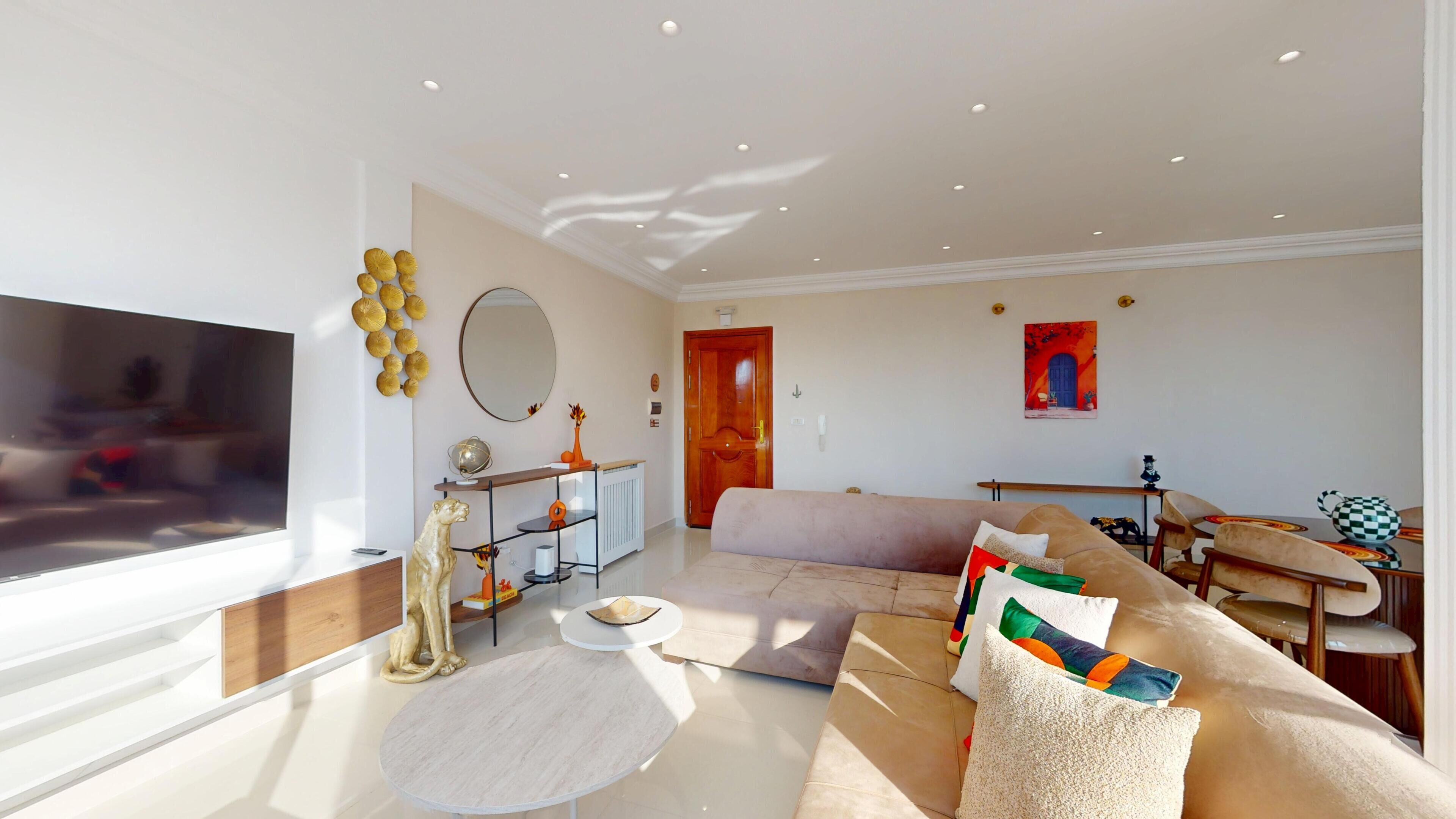Una sala de estar moderna con un televisor de pantalla plana grande, un sofá, una mesa de centro y un comedor con una mesa y sillas.