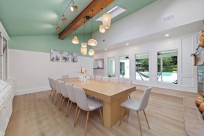 Un vaste espace de salle à manger avec une longue table en bois et des chaises blanches, une cuisine avec des meubles blancs et une vue sur une piscine, visible à travers de grandes fenêtres.