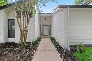 A narrow pathway leading to a white building with a black-framed door, surrounded by a garden with various plants and a tree. - Lara Retreat- 6B/4.5 BT- Private Pool and Paris- Game room (Houston)