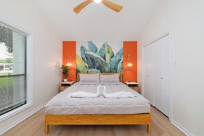 Quarto com cama, ventoinha no teto, pintura na parede, abajures e janela com persianas.