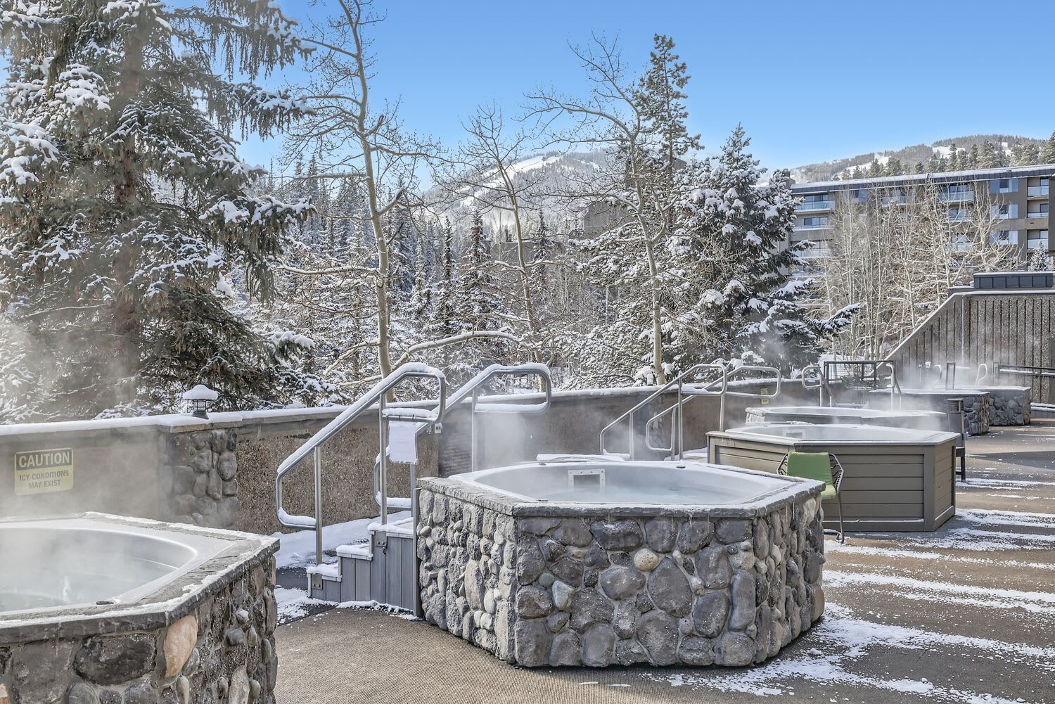 A snow-covered outdoor area with hot tubs and stone walls, surrounded by trees and buildings.