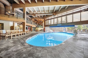 Una piscina interior con un cartel que indica ‘no saltos’, bancos de madera y vistas al exterior a través de amplios ventanales.