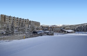 Estación de esquí nevada con un gran edificio de hotel, un paseo techado y montañas al fondo.