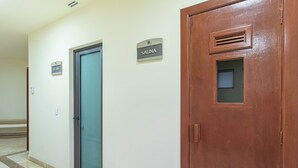 En hall med dörrar märkta med ”BASTA” och ”ÅNGBASTU”, en trädörr med ett ventilationsgaller och ett klinkergolv.