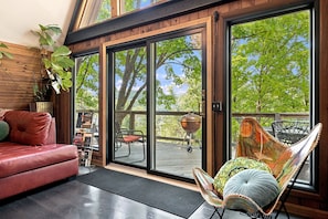 A modern living room with a leather sofa, a cushioned chair, and a large sliding glass door leading to an outdoor deck with a barbecue grill.