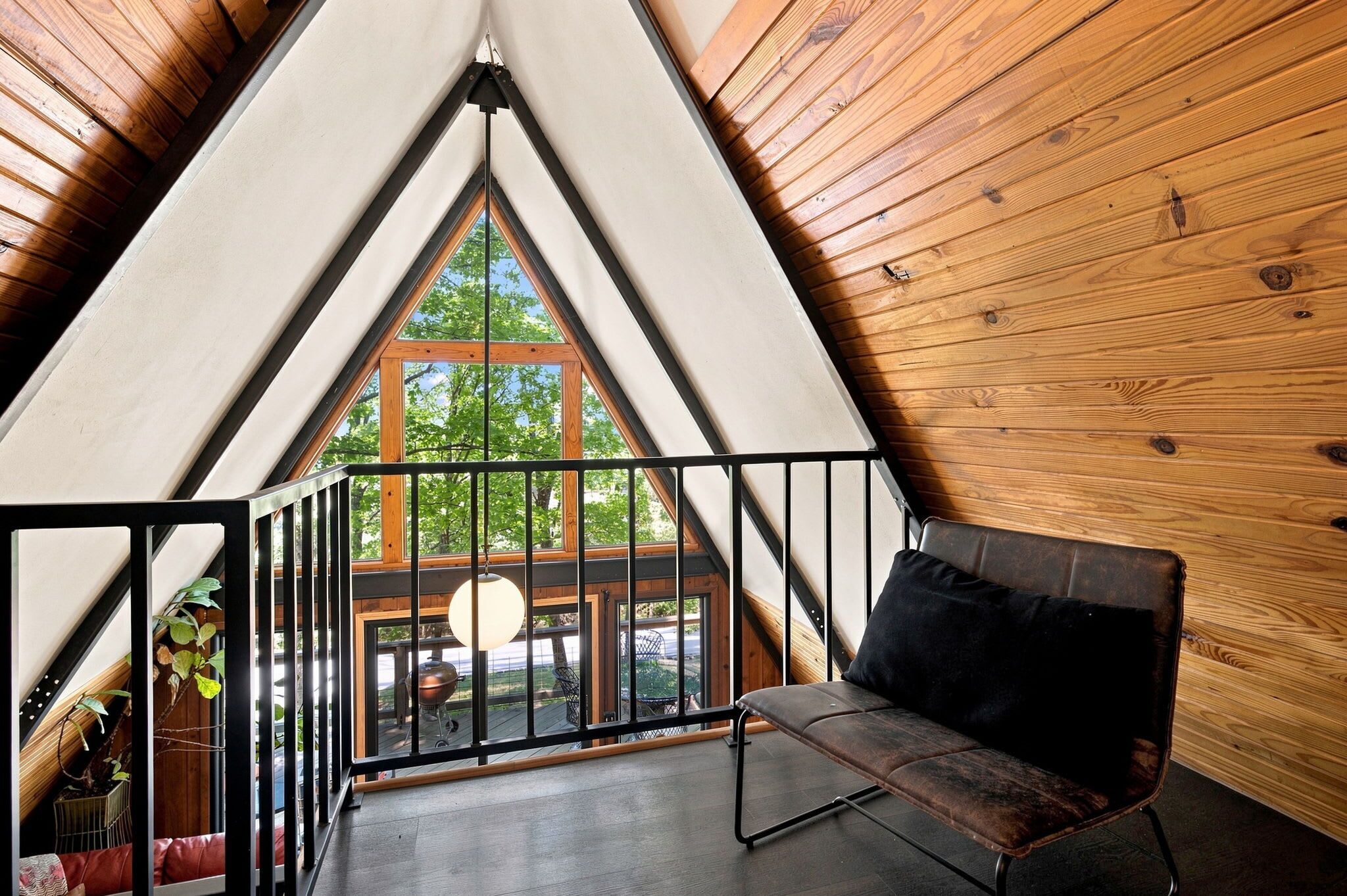 A cozy attic room with a sloped ceiling, a black metal railing, a cushioned bench, and a view of a backyard with a barbecue grill.