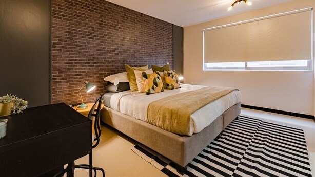 A modern bedroom with a brick accent wall, a large bed with patterned pillows, a desk with a lamp, and a large window with blinds. - Two Roma Norte Condos with City Views & Workspace (Ciudad de México)
