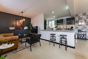 A modern living room with a sofa, a coffee table, and a TV. There is a kitchen area with a bar and stools. - Two Roma Norte Condos with City Views & Workspace (Ciudad de México)
