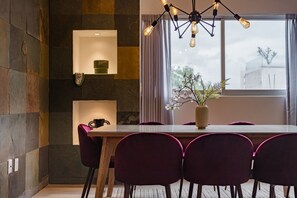 A modern dining area with a wooden table, purple chairs, a vase with flowers, and a unique light fixture. - Two Roma Norte Condos with City Views & Workspace (Ciudad de México)