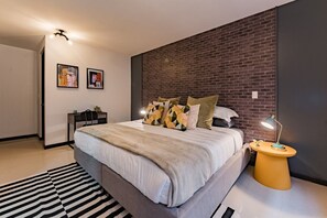 Een moderne slaapkamer met een metselwerk accentmuur, een groot bed met veel kussens, een bureau en een staande lamp.