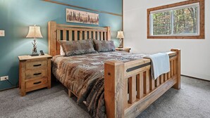 Un dormitorio con una cama de madera, dos mesitas de noche, una ventana con vistas a árboles y una pared con un cuadro.