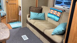 A compact RV interior with a kitchenette, a sofa with cushions, a clock, and a window with an 'EXIT' sign.