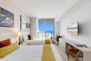 A hotel room with two beds, a desk, a TV, and a balcony with a city view. - 2BR Ocean Views & Modern Comfort (Hollywood)