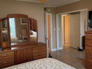 A bedroom with a wooden dresser, a bed with a patterned bedspread, a full-length mirror, and a door leading to another room. - Beachfront Condo - 90-Night Minimum Stay (Clearwater)