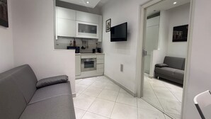 Ein moderner Wohnraum mit grauem Sofa, einer Küchenzeile mit weißen Schränken, einem Flachbildfernseher und einem grauen Schlafsofa in einem separaten Raum.