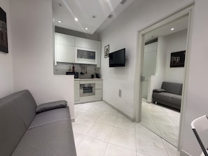 A modern living space with a grey sofa, a kitchen area with white cabinets, a flat-screen TV, and a grey couch in a separate room. - Sisto Suite - Charming 1-bedroom apartment with AC in vibrant Roma (Roma)