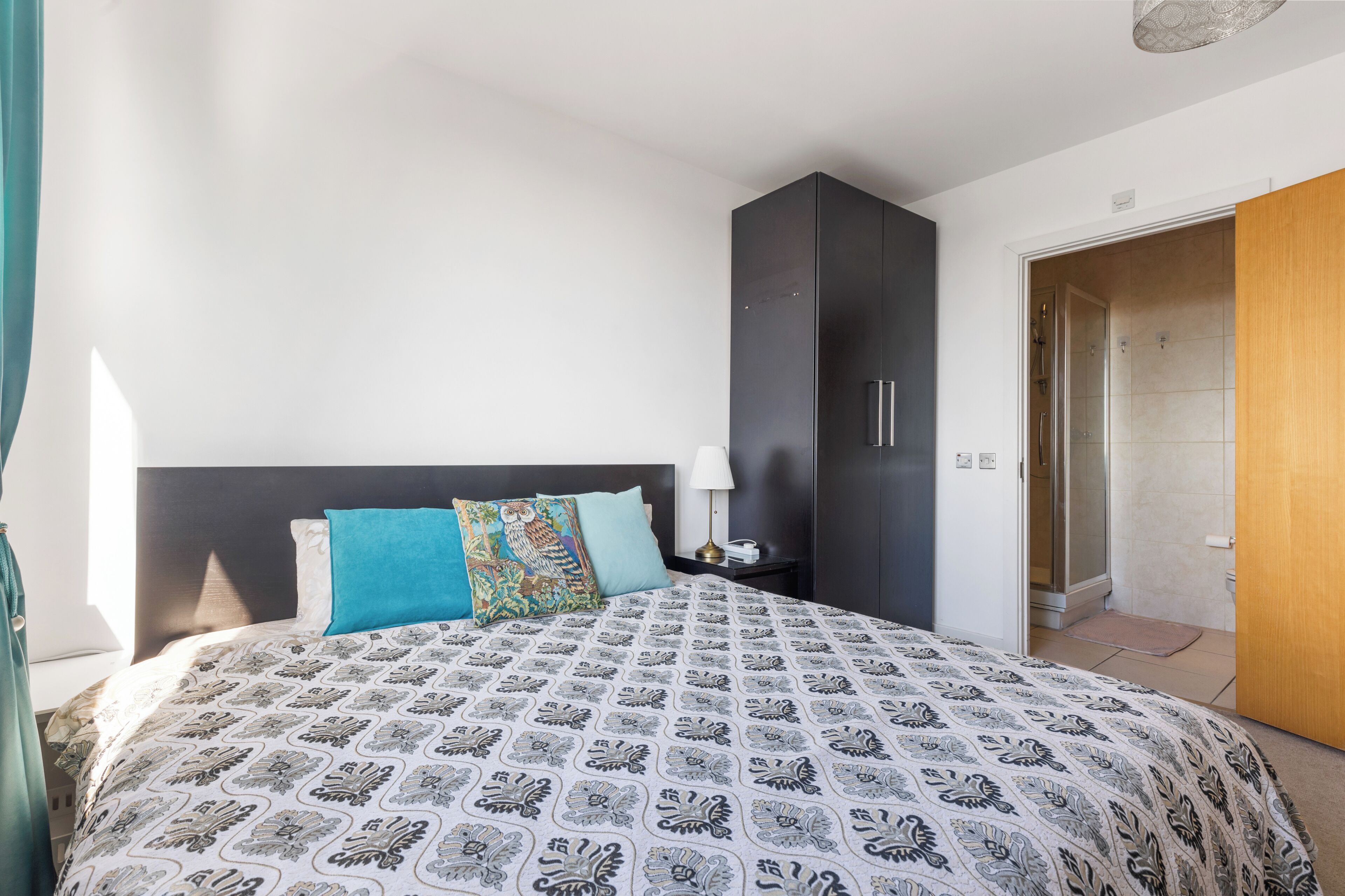 A bedroom with a bed, a nightstand, a lamp, a wardrobe, and a bathroom door.