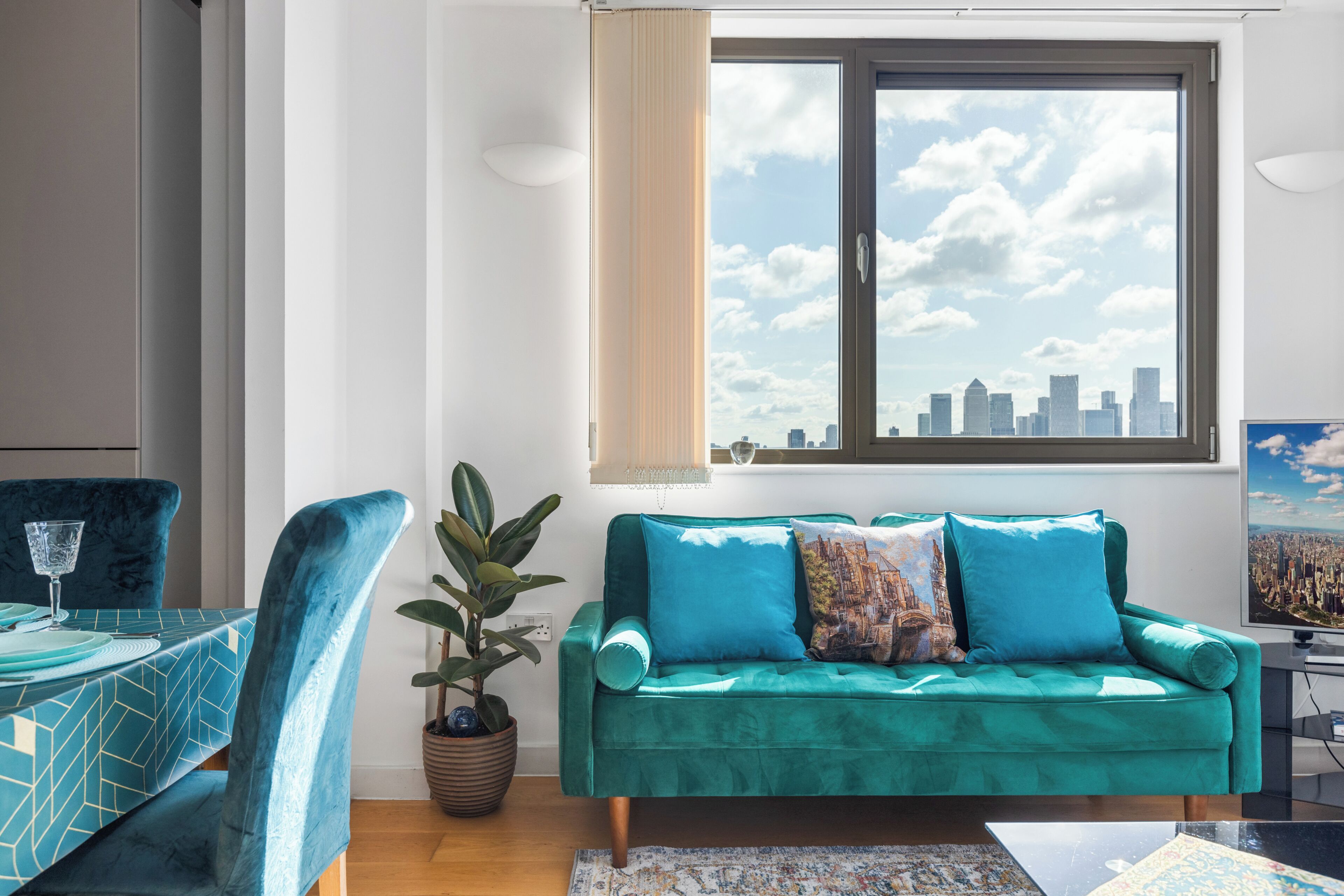 Ein Wohnzimmer mit einem petrolfarbenen Sofa, einem Esstisch mit einem Glas und Blick auf die Stadt durch ein Fenster.