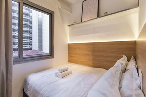 Una habitación de hotel con cama, ventana con vista a edificios y repisa con obras de arte enmarcadas.