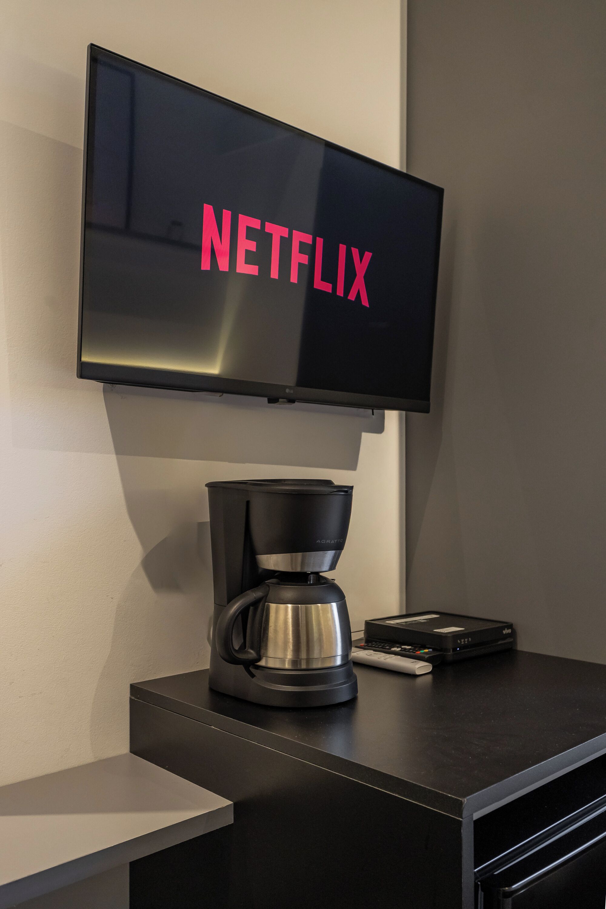 A flat-screen TV mounted on a wall with the word 'NETFLIX' in pink letters. Below the TV, there is a black coffee maker and a remote control on a black surface.