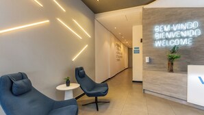 Ein moderner Empfangsbereich mit einem leuchtenden Neon-Schild, zwei blauen Stühlen, einem kleinen Tisch mit Pflanze und einer Wand mit Hinweisschildern.