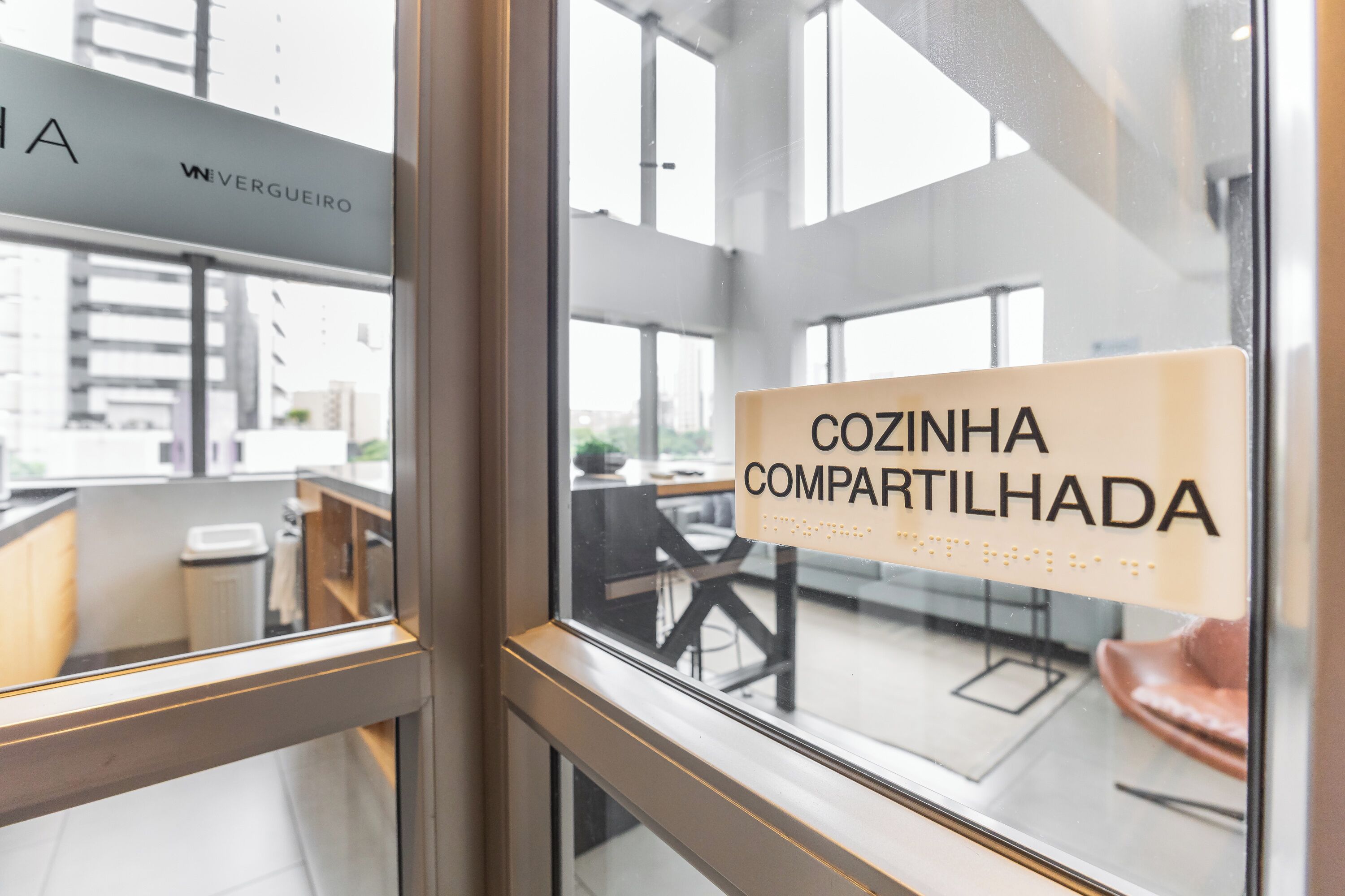 Une fenêtre sur laquelle on peut lire l’inscription « COZINHA COMPARTILHADA » et, à travers laquelle, on aperçoit un espace de bureau avec un bureau et une chaise.