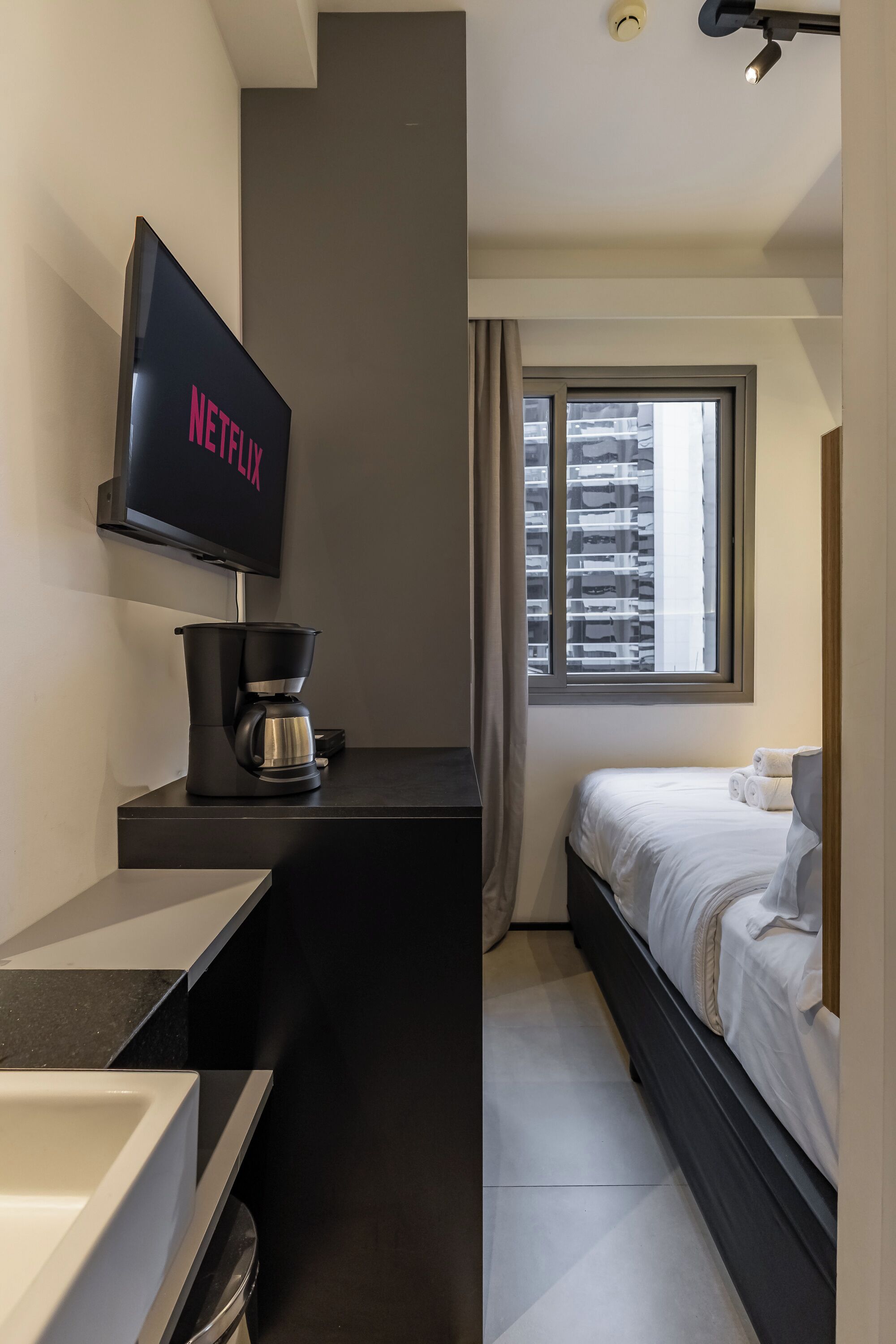 Habitación de hotel moderna con televisor de pantalla plana en la pared, cafetera en la encimera, cama con ropa blanca y ventana con persianas.