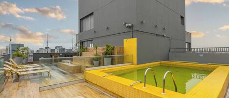 A rooftop pool area with a modern building in the background, a glass balustrade, and lounge chairs.